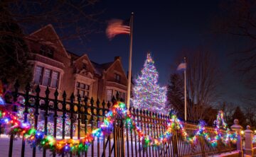 multi-colored holiday lights draped along fence and wrapped around large conifer in front of governor's mansion