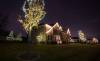 Two story home with white lights on roof line and several small trees wrapped with white Christmas lights.